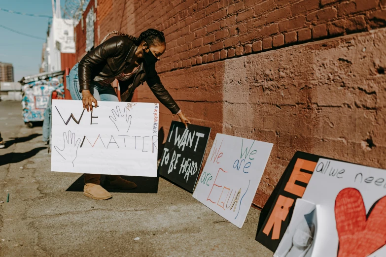 a man leaning against a brick wall holding a sign, a photo, by Julia Pishtar, trending on pexels, black arts movement, protesters holding placards, kneeling, thumbnail, kailee mandel