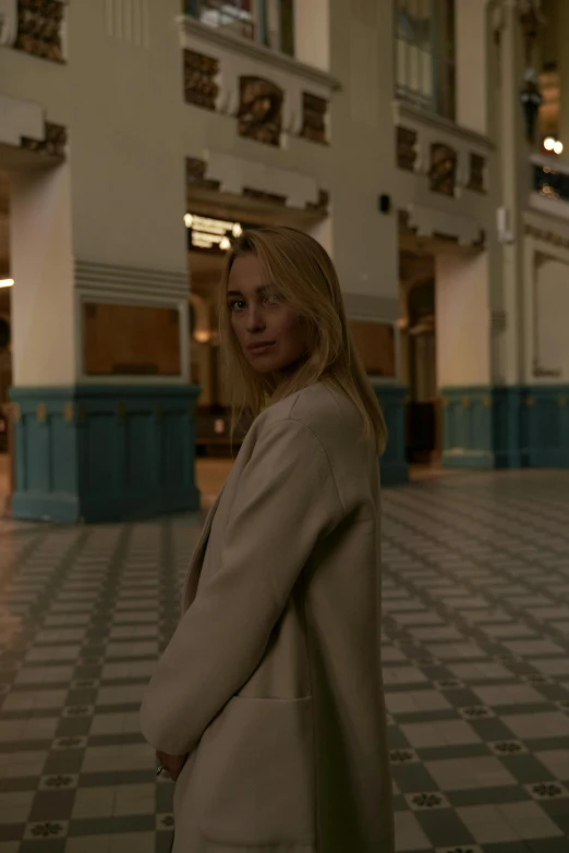 a woman is standing in a large building, inspired by Henri-Julien Dumont, tv show still, she wears a jacket, ignant, blonde woman