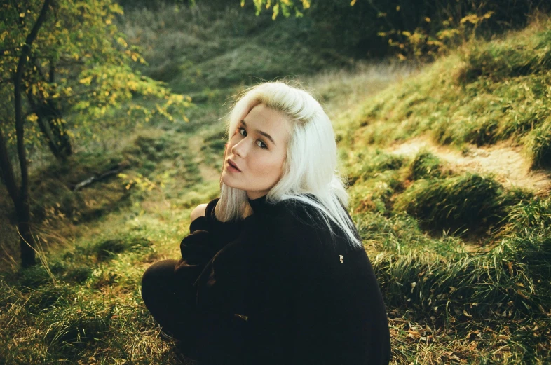 a woman sitting on top of a lush green hillside, an album cover, inspired by Elsa Bleda, pexels contest winner, white-hair pretty face, profile image, chilly dark mood, looking towards camera