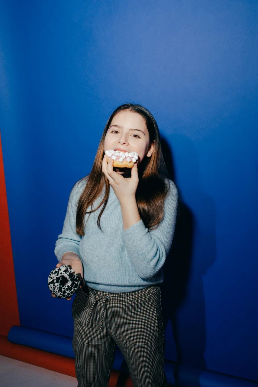 a woman standing in front of a blue wall eating a donut, a polaroid photo, dilraba dilmurat, at a birthday party, holding toasted brioche bun, in front of a black background