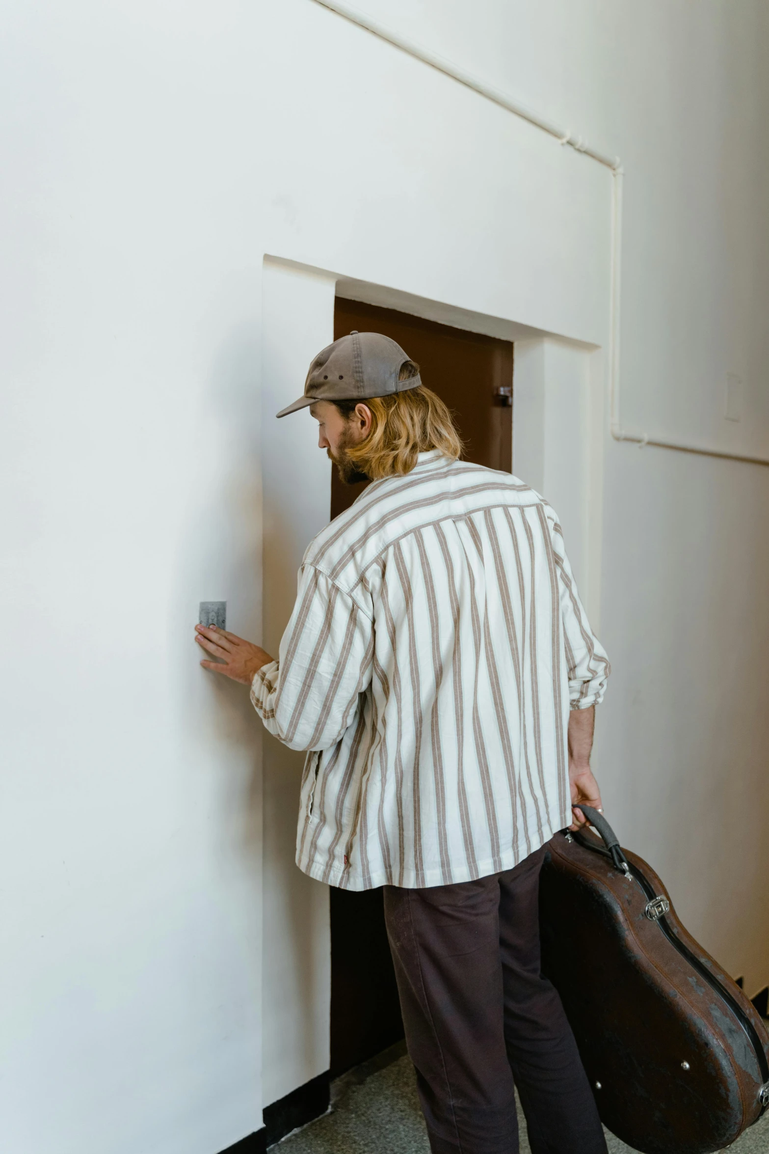 a man standing next to a wall with a guitar case, pexels contest winner, happening, wearing stripe shirt, leaving a room, caracter with brown hat, about to enter doorframe