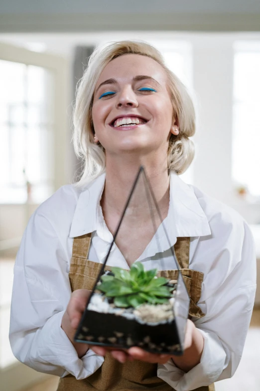 a woman holding a plant in a glass container, a colorized photo, pexels contest winner, aestheticism, earing a shirt laughing, white apron, blonde swedish woman, happy female alien