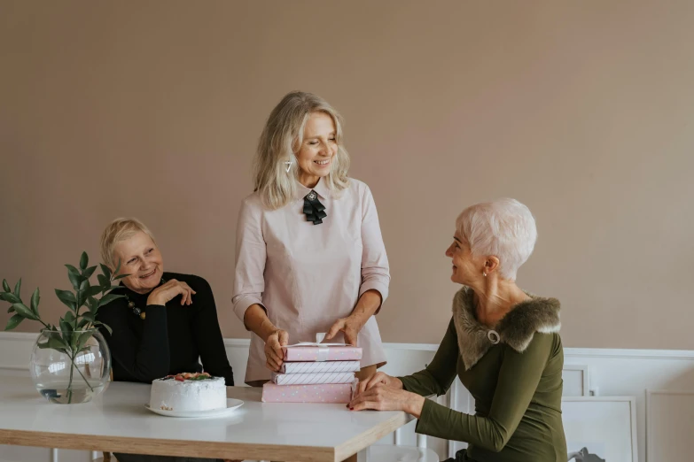 two women sitting at a table with a cake in front of them, by Emma Andijewska, pexels contest winner, white haired lady, three women, standing elegantly, on a white table