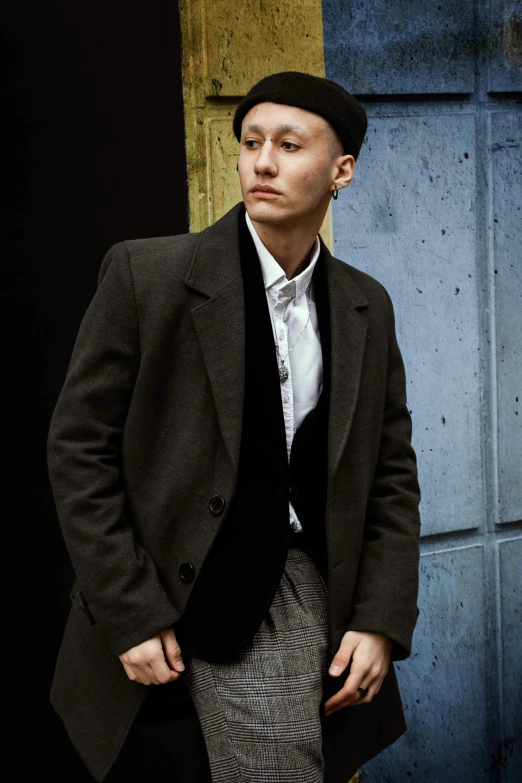 a man in a suit and hat standing in front of a wall, an album cover, inspired by Yukihiko Yasuda, unsplash, wearing a turtleneck and jacket, androgynous person, actor, early evening