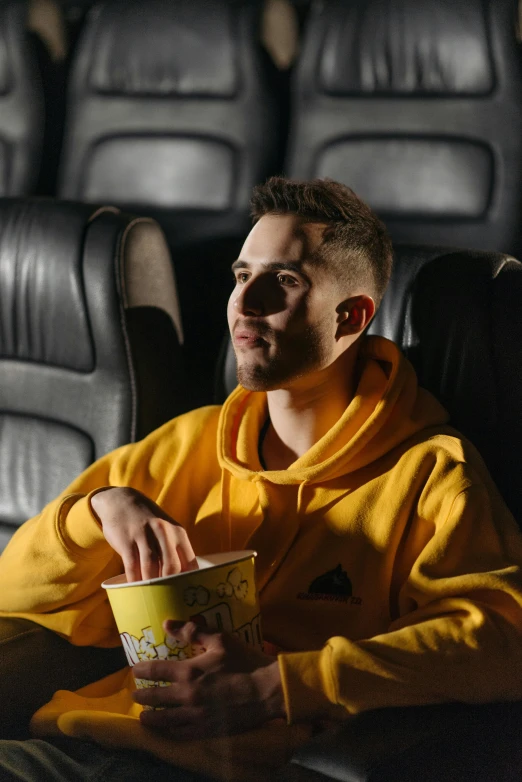 a man sitting in a chair with a cup of popcorn, a portrait, trending on pexels, wearing a yellow hoodie, avatar image, ( ( theatrical ) ), a handsome