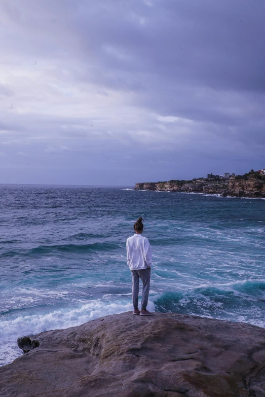 a man standing on top of a rock next to the ocean, a picture, inspired by Sydney Carline, trending on unsplash, she's sad, bondi beach in the background, standing in a maelstrom, standing in a grotto
