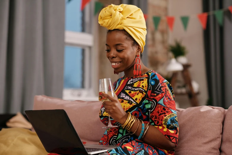 a woman sitting on a couch using a laptop, inspired by Chinwe Chukwuogo-Roy, pexels, afrofuturism, enjoying a glass of wine, wearing festive clothing, cloth head wrap, colourful clothes