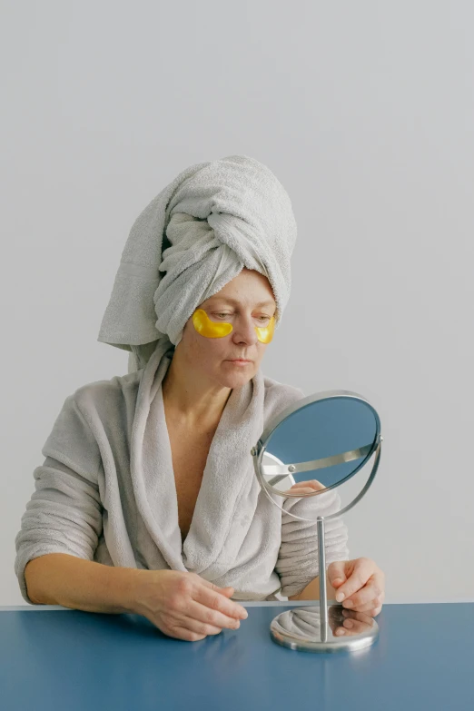 a woman sitting at a table with a towel on her head, with a mirror, turning yellow, mirror eye implants, grey