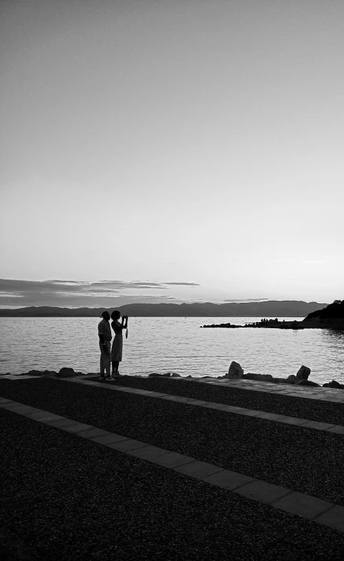 a couple of people standing next to a body of water, a black and white photo, by Alison Watt, sunset!!!, photo session, split near the left, romantic!!!