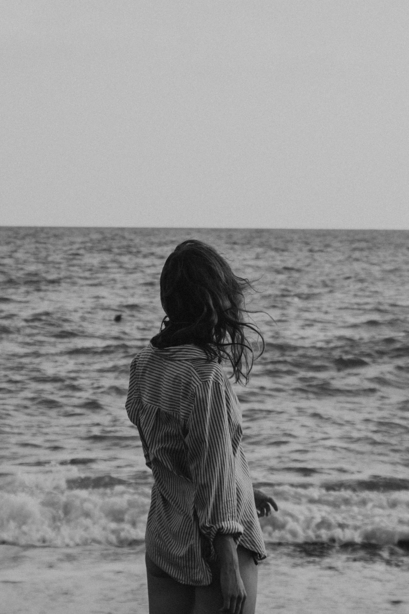 a woman standing on top of a beach next to the ocean, a black and white photo, the sea of sadness, (beautiful) girl, chillwave, with his back turned