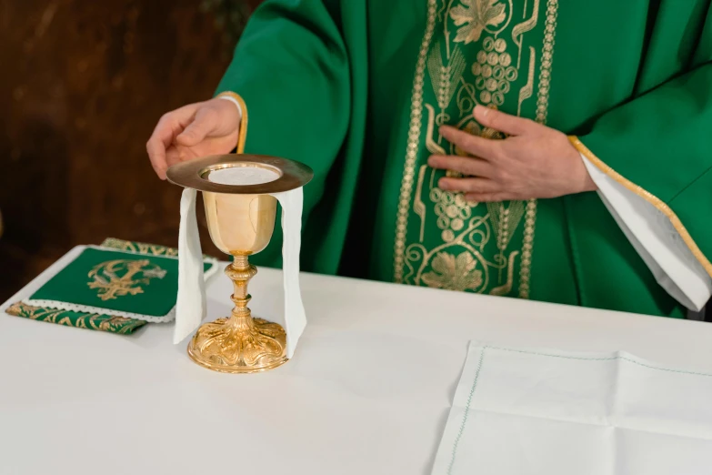 a priest holding a golden cup on top of a table, pexels, green litham veil, neoprene, serving body, panels