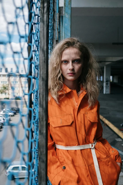 a woman in an orange coat leaning against a fence, inspired by Elsa Bleda, pexels contest winner, wearing prison jumpsuit, worksafe. instagram photo, ellie bamber, photo of a model