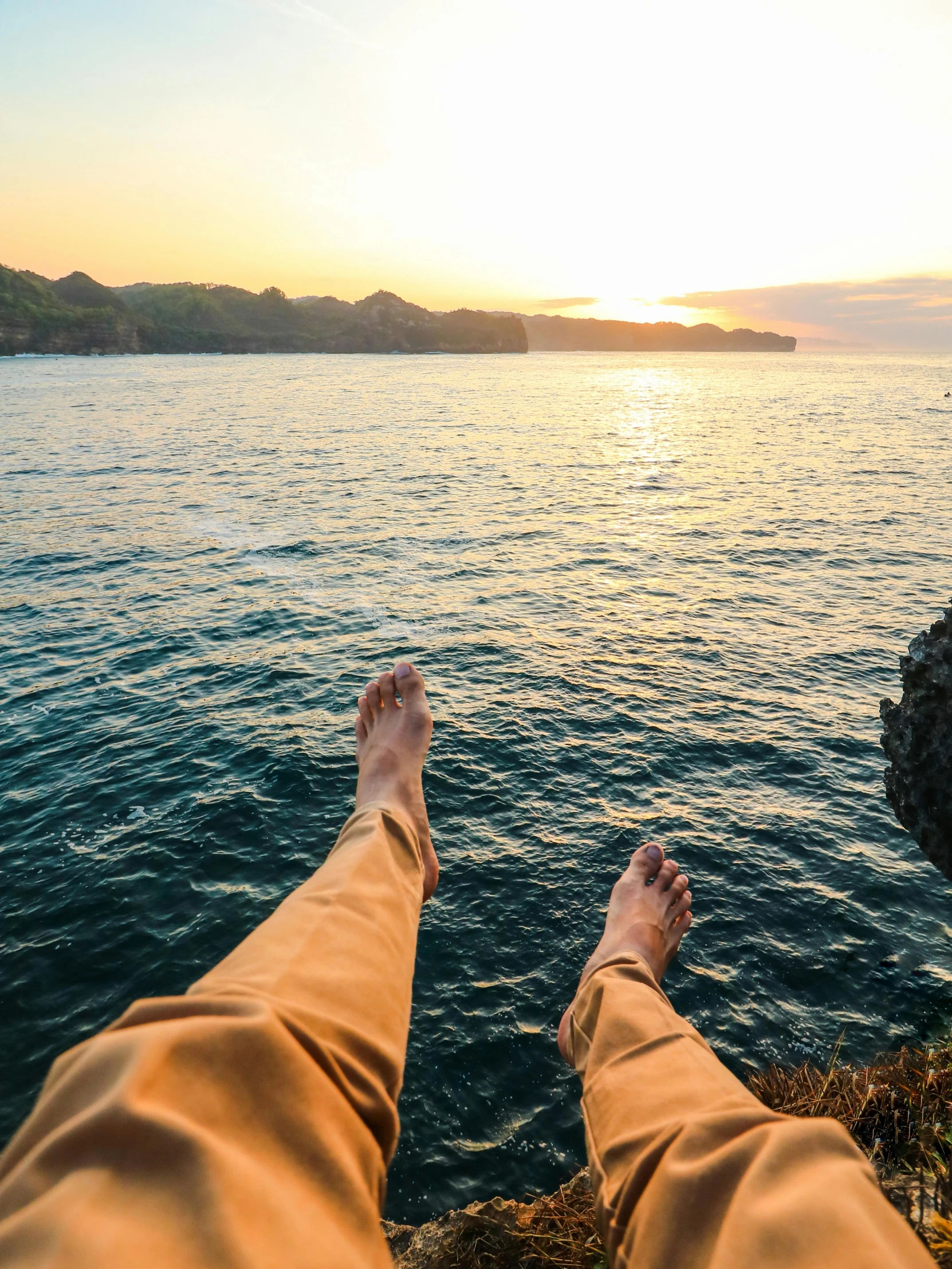 a person sitting on top of a rock next to a body of water, barefeet, during a sunset, full body pov, steep cliffs