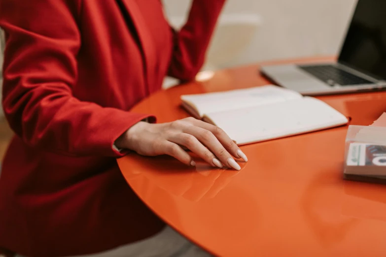 a woman sitting at a table in front of a laptop computer, by Emma Andijewska, trending on pexels, private press, red suit, white and orange, diary on her hand, immaculate detail