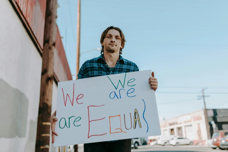 a man holding a sign that says we are equal, by Julia Pishtar, trending on pexels, renaissance, square, carson ellis, frank dillane, color picture