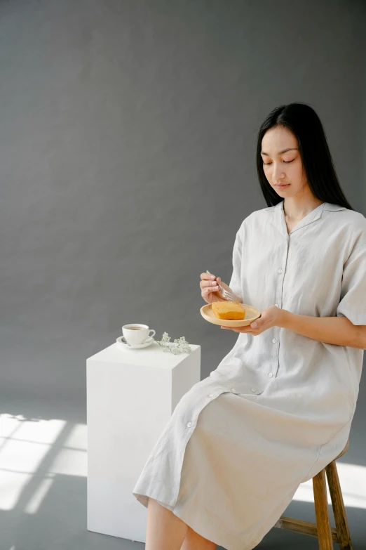 a woman sitting on a stool holding a plate of food, inspired by Li Di, trending on unsplash, minimalism, soft silk dress, grey, honey, low quality photo