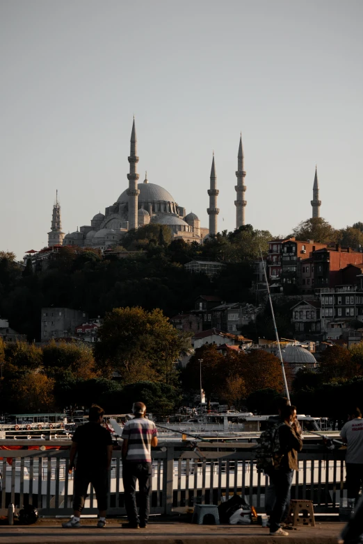 a group of people standing on top of a bridge, a picture, inspired by Altoon Sultan, pexels contest winner, hurufiyya, black domes and spires, viewed from the harbor, in the hillside, late summer evening