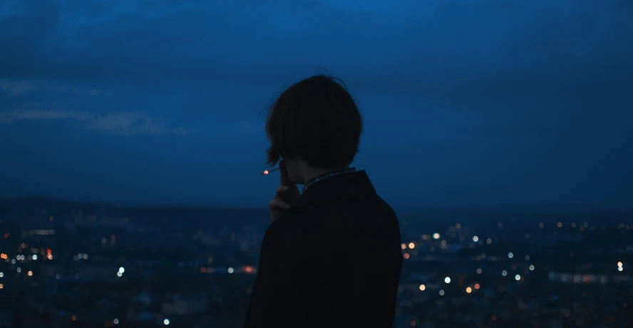 a person standing in front of a city at night, pexels contest winner, ☁🌪🌙👩🏾, human staring blankly ahead, deep blue mood, teenage boy