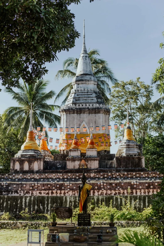 a large white and gold building surrounded by trees, a statue, unsplash contest winner, laos, prayer flags, coconut trees, old ruins