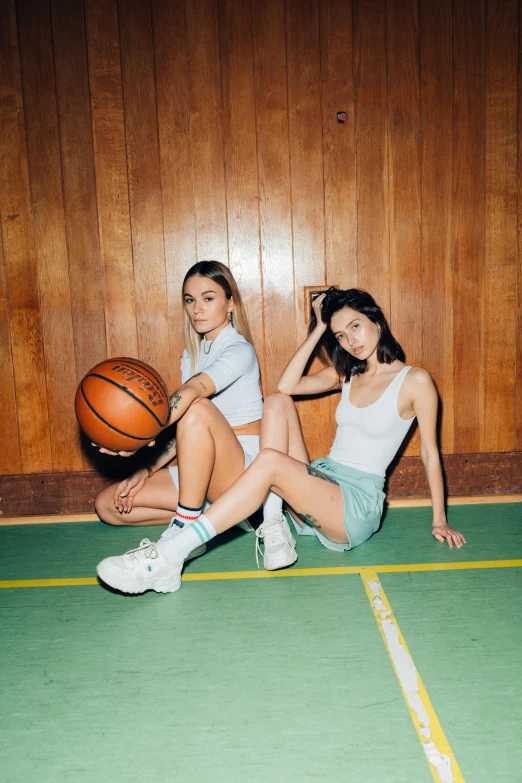 a couple of women sitting on top of a basketball court, an album cover, wearing white sneakers, sitting on a mocha-colored table, sports setting, gemma chen