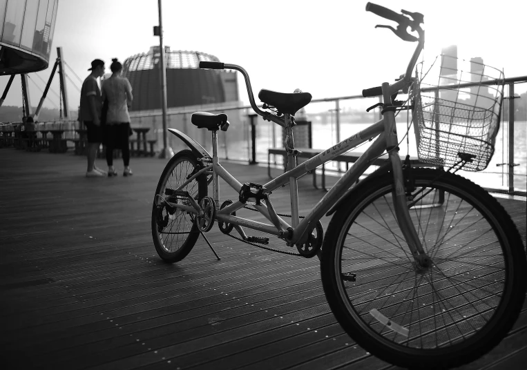 a black and white photo of a bicycle on a pier, people watching around, good times photograph