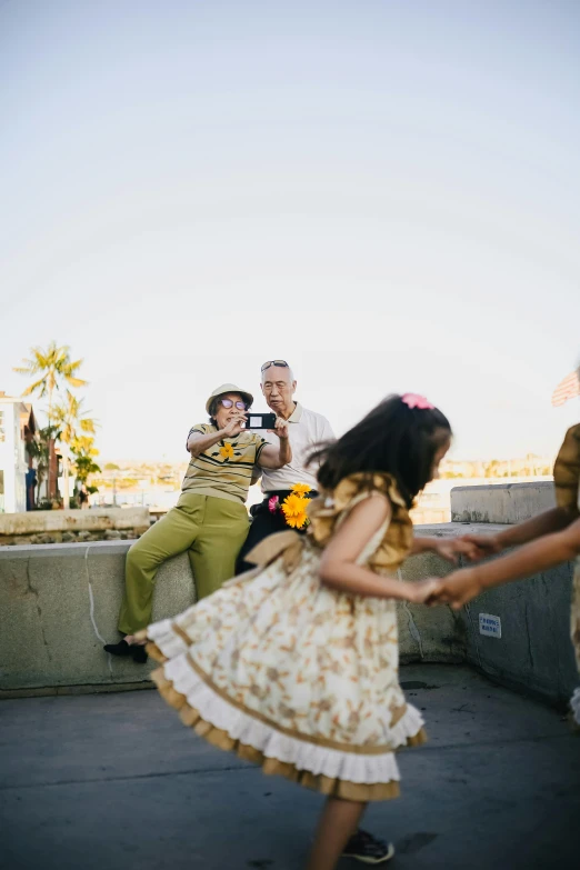 a group of people sitting on top of a cement wall, an album cover, inspired by Norman Rockwell, unsplash, happening, wearing yellow floral blouse, dancing a jig, los angeles 2 0 1 5, families playing