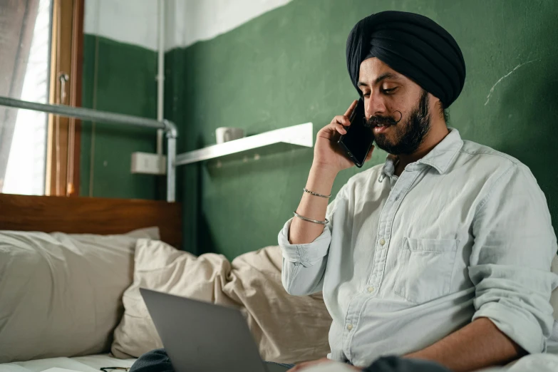 a man sitting on a bed talking on a cell phone, inspired by Manjit Bawa, trending on pexels, renaissance, sitting at a computer, aboriginal australian hipster, turban, thumbnail