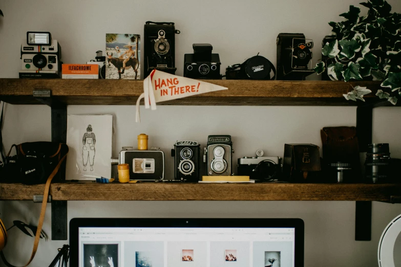 a laptop computer sitting on top of a wooden desk, a picture, by Carey Morris, trending on pexels, maximalism, jar on a shelf, vintage camera, shelves, lovingly looking at camera