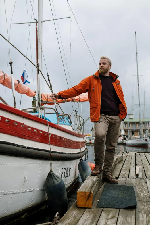 a man standing on a dock next to a boat, inspired by Jóhannes Sveinsson Kjarval, happening, orange jacket, he's on an old sailing boat, lgbtq, reykjavik