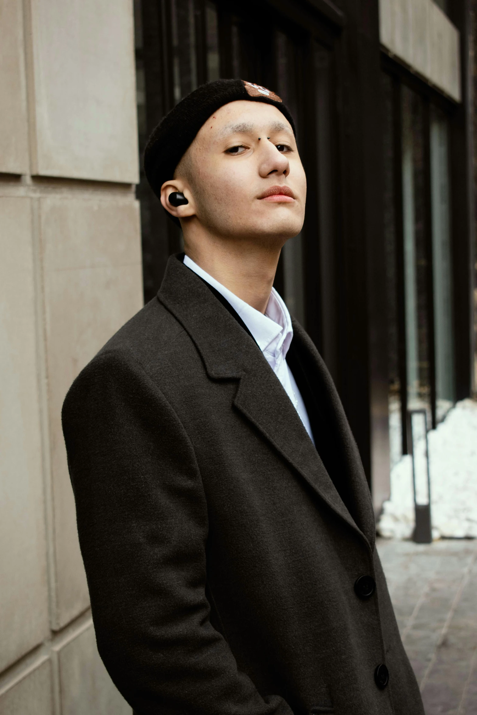 a man in a black coat standing in front of a building, inspired by Chen Chi, trending on pexels, renaissance, earbuds jewelry, non binary model, left ear, small chin