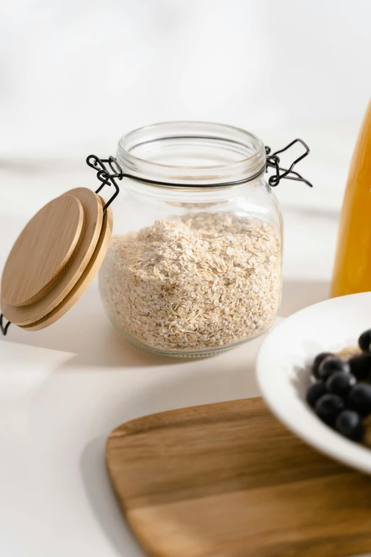 a white plate topped with blueberries next to a jar of oatmeal, a picture, unsplash, renaissance, glassware, yeast, detail shot, a wooden