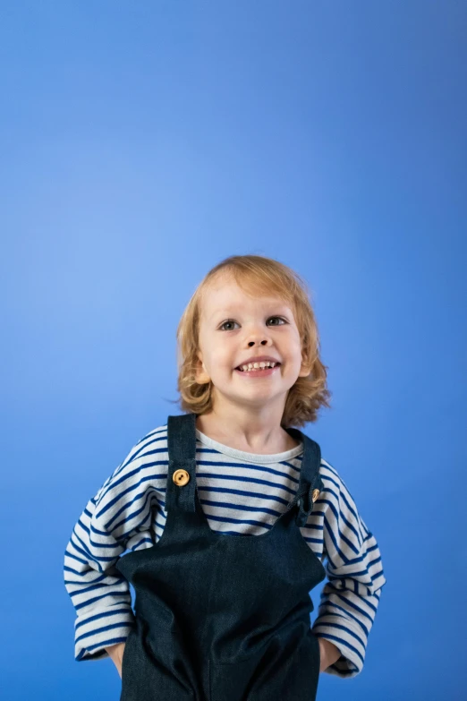 a little boy standing in front of a blue background, while smiling for a photograph, product introduction photos, wearing overalls, wearing stripe shirt