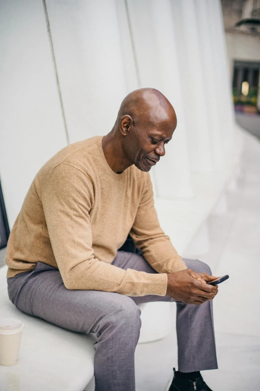 a man sitting on a bench looking at his cell phone, brown sweater, emmanuel shiru, mobile learning app prototype, mature male
