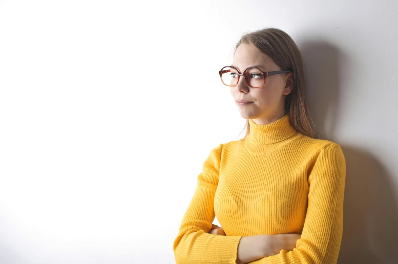 a woman wearing glasses leaning against a wall, trending on pexels, wearing turtleneck, yellow, on white background, a lonely woman