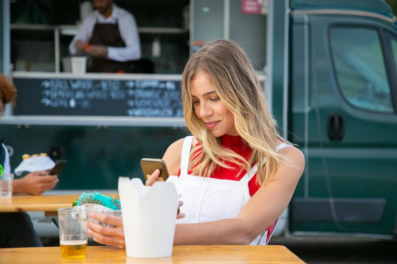 a woman sitting at a table in front of a food truck, by Julian Allen, pexels, goddess checking her phone, square, sydney sweeney, wearing an apron