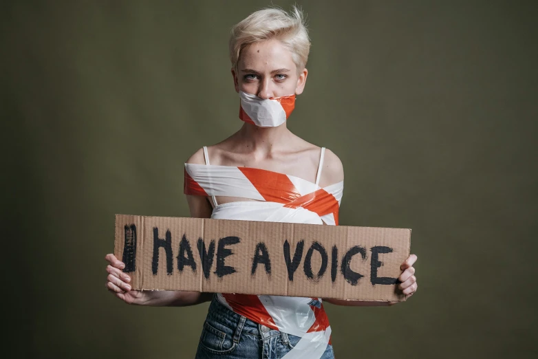 a woman holding a cardboard sign that says i have a voice, pexels, white and orange breastplate, young blonde woman, bandage, no stipe