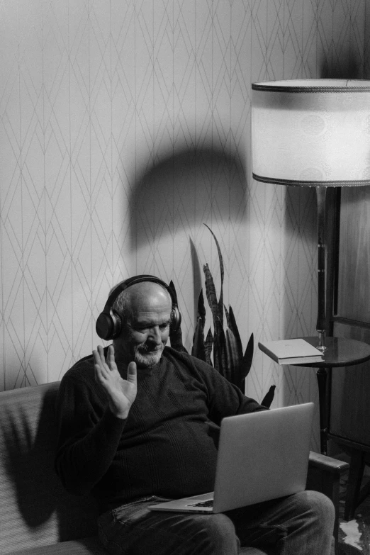 a man sitting on a couch using a laptop computer, a black and white photo, inspired by Arnold Newman, with head phones, old gigachad with grey beard, waving, mike ehrmantraut