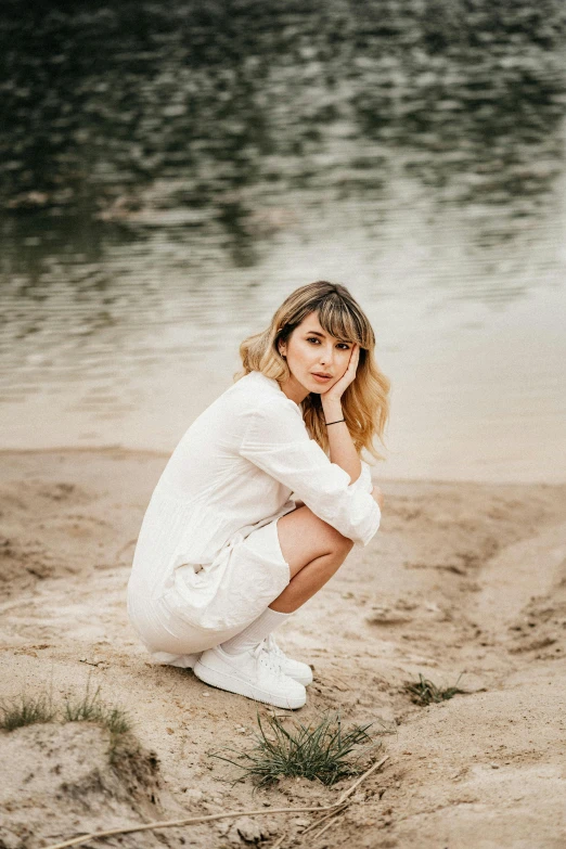 a woman sitting on a beach next to a body of water, an album cover, trending on pexels, in white clothes, portrait pose, whitebangs, ekaterina