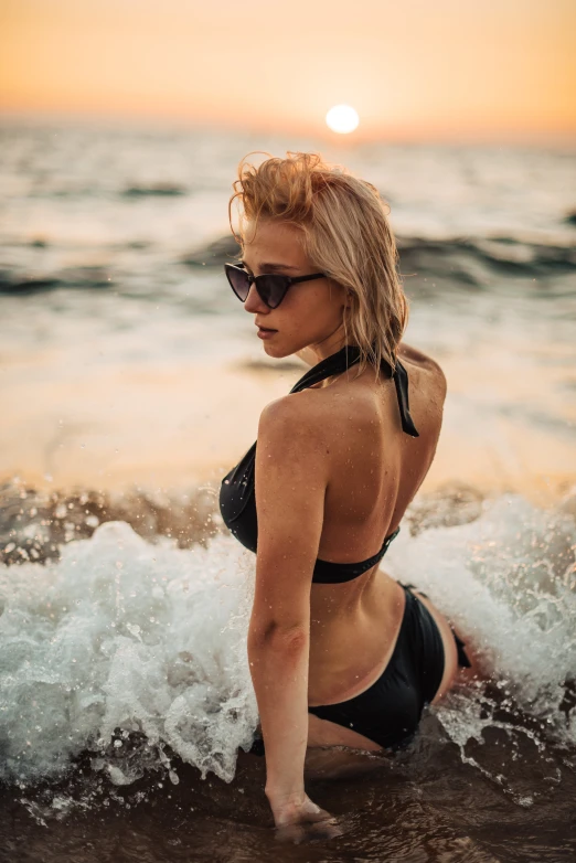 a woman sitting on top of a beach next to the ocean, black swimsuit, crashing waves, implanted sunglasses, close up of a blonde woman