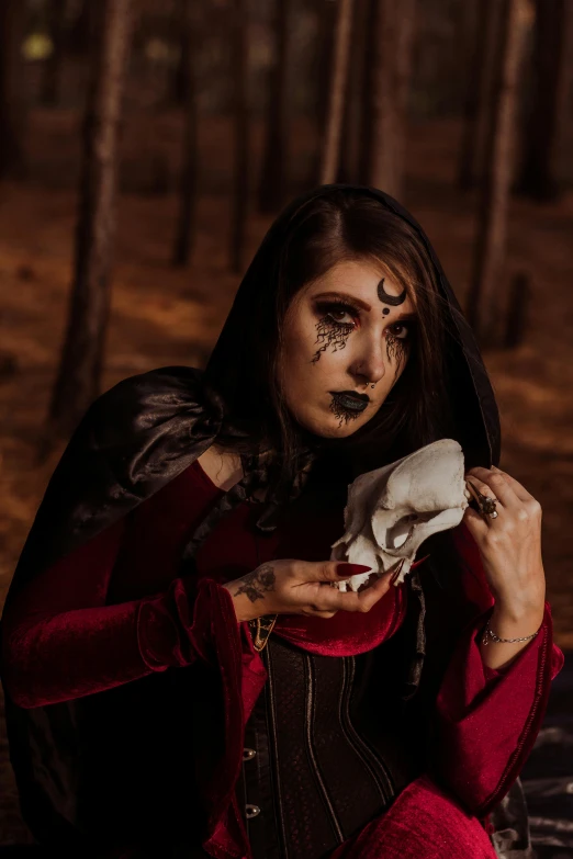 a woman dressed as a witch sitting in the woods, an album cover, inspired by Samuel Hieronymus Grimm, pexels contest winner, gothic art, holding a skull, profile image, wearing cultist red robe, corpsepaint