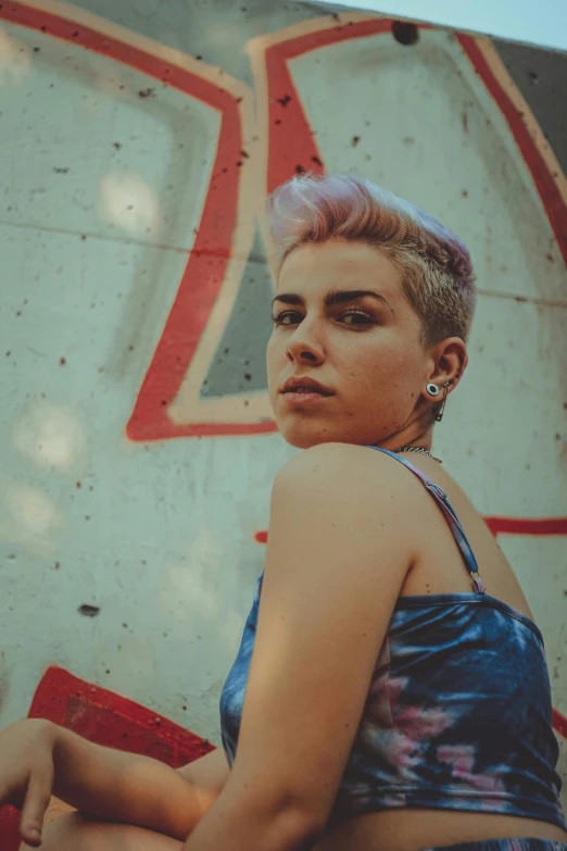 a woman sitting in front of a graffiti covered wall, an album cover, inspired by Elsa Bleda, trending on pexels, short purple hair with undercut, cinestill 800t eastmancolor, headshot, androgynous male