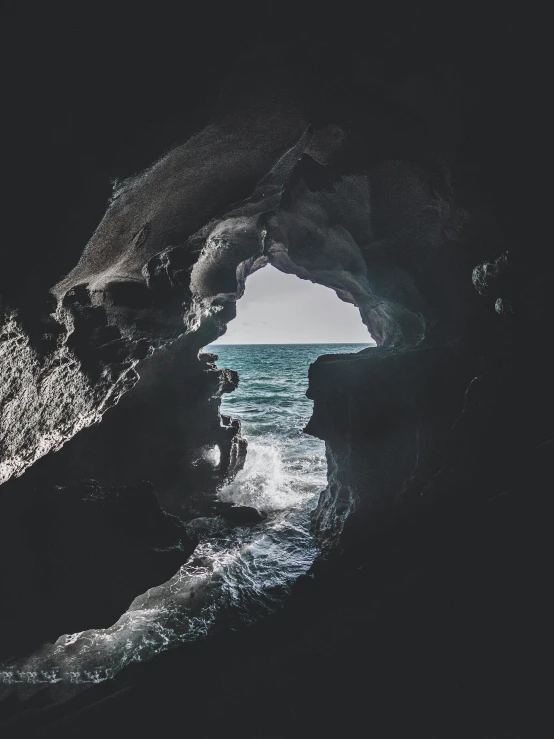 a man standing inside of a cave next to the ocean, dark and white, devils horns, inside the curl of a wave, through the windows