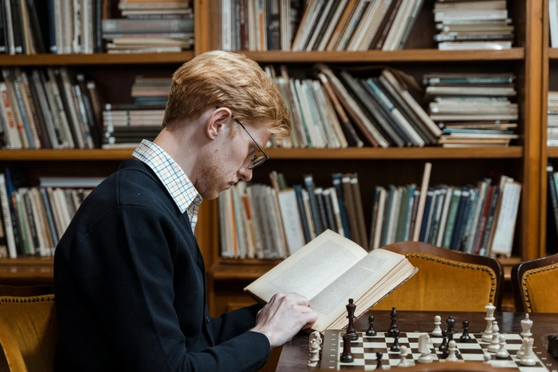 a man sitting at a table reading a book, inspired by Simon Marmion, pexels contest winner, academic art, chess set, looks like domhnall gleeson, library books, thumbnail