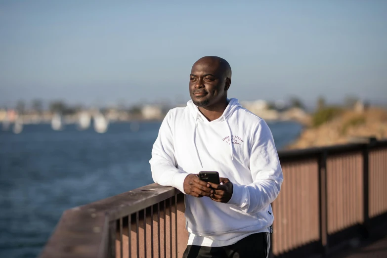 a man standing on a bridge looking at his cell phone, huell babineaux, profile image, wearing a track suit, ocean in the background