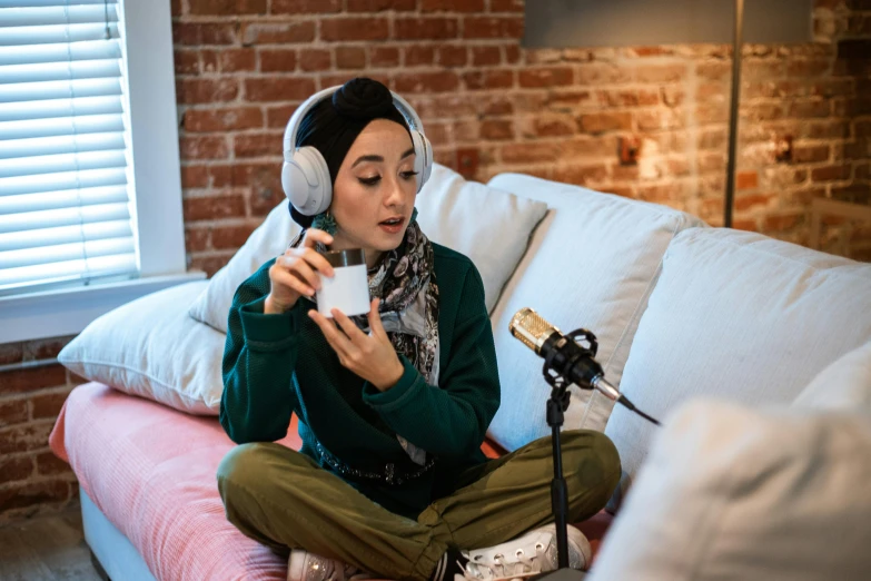 a woman sitting on a couch listening to music, a portrait, trending on pexels, hurufiyya, white hijab, table with microphones, drinking coffee at central perk, model wears a puffer jacket