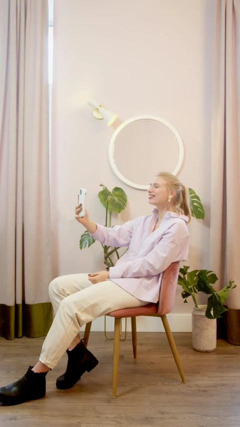 a woman sitting in a chair holding a remote control, by Paul Bird, pexels contest winner, round mirror on the wall, taking a selfie, pastel lighting, next to a plant