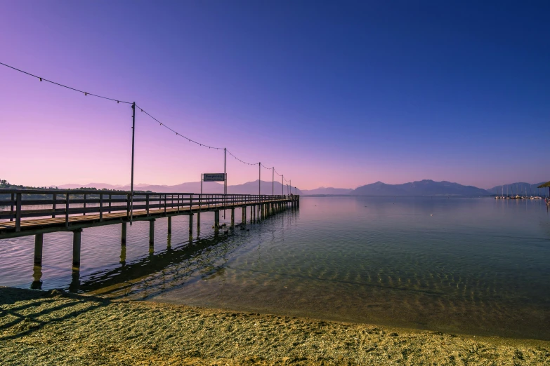 a pier in the middle of a body of water, by Niko Henrichon, distant mountains lights photo, clear blue skies, today\'s featured photograph 4k, sunset beach