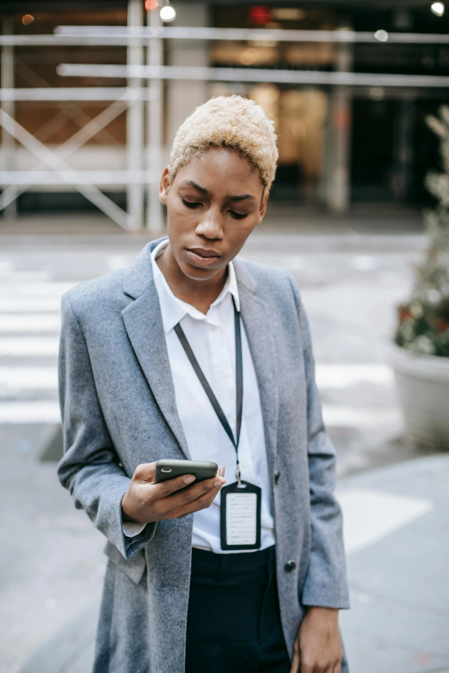 a woman standing on a sidewalk looking at her cell phone, trending on pexels, renaissance, wearing black grey suit, short blonde afro, security agent, non-binary