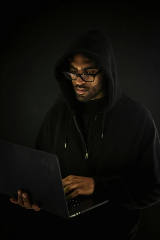 a man in a black hoodie using a laptop, dark-skinned, dark and forboding, serious lighting, dark. no text