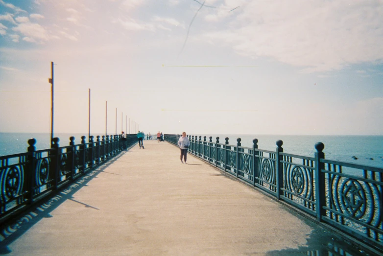 a person walking on a pier next to the ocean, a polaroid photo, unsplash, blue tint expired film, bridges and railings, vhs colour photography, no watermarks